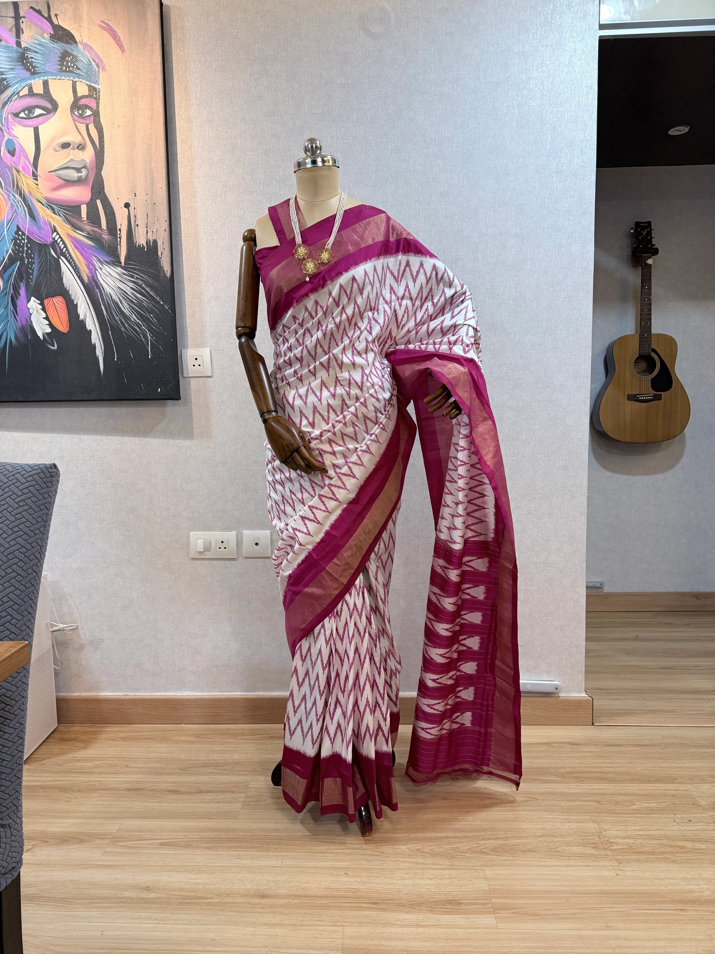 Maroon-and-white chevron ikat saree on the mannequin, pallu draped over the shoulder with gold zari border, brass jewellery visible at the neckline.