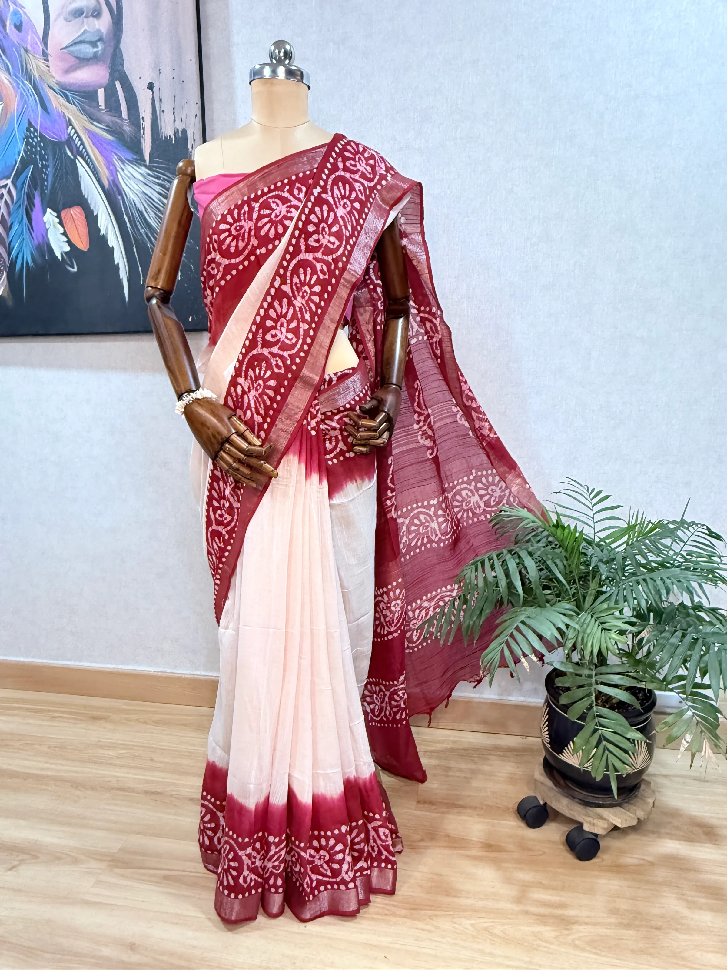 Maroon batik-print linen saree draped on a mannequin, pallu spread out beside a potted palm, showing the block-print floral border on cream ground.