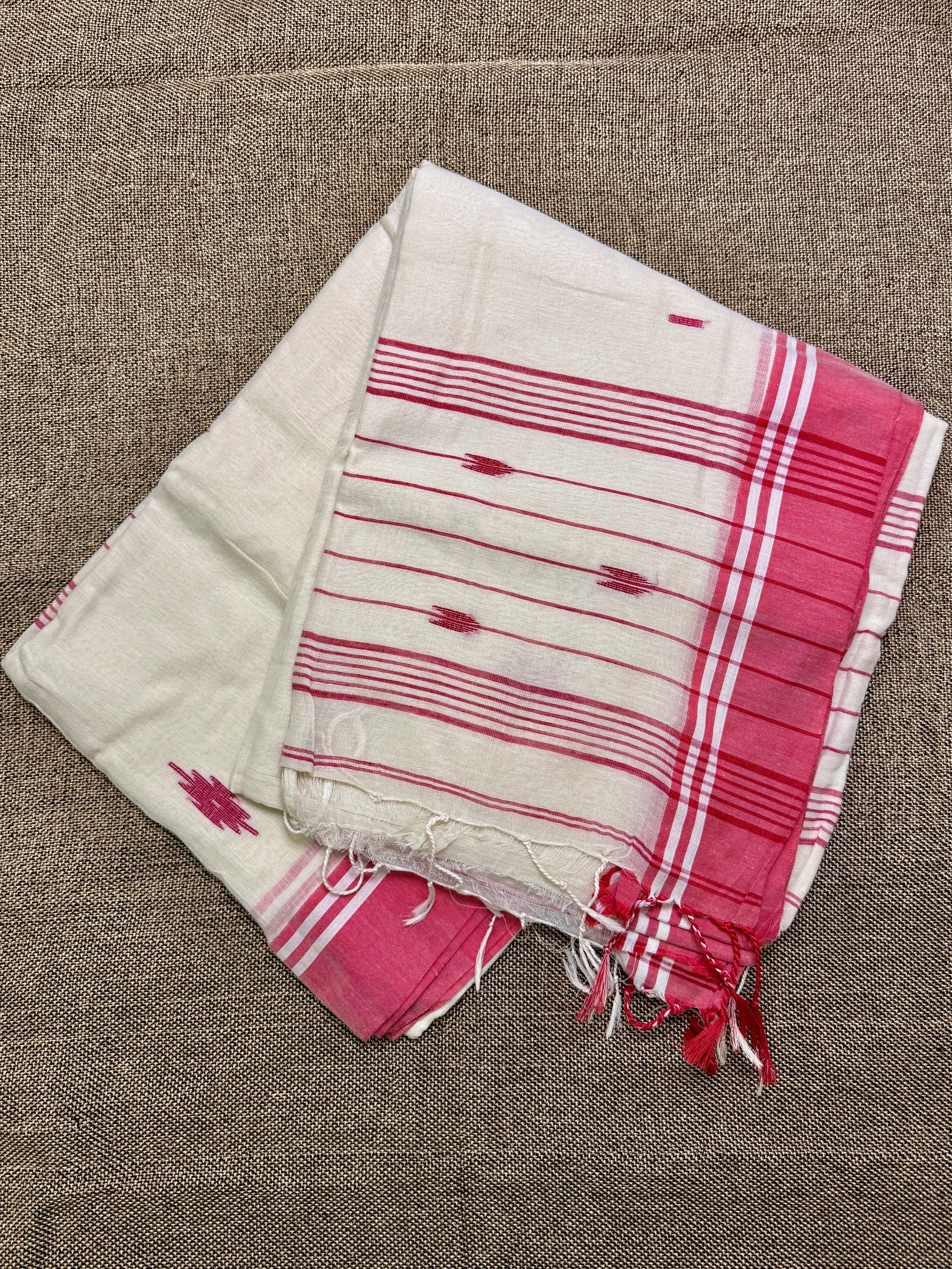 Folded cream mul cotton Tangail saree with fine red stripes and a pink plaid border, laid on a woven jute surface.