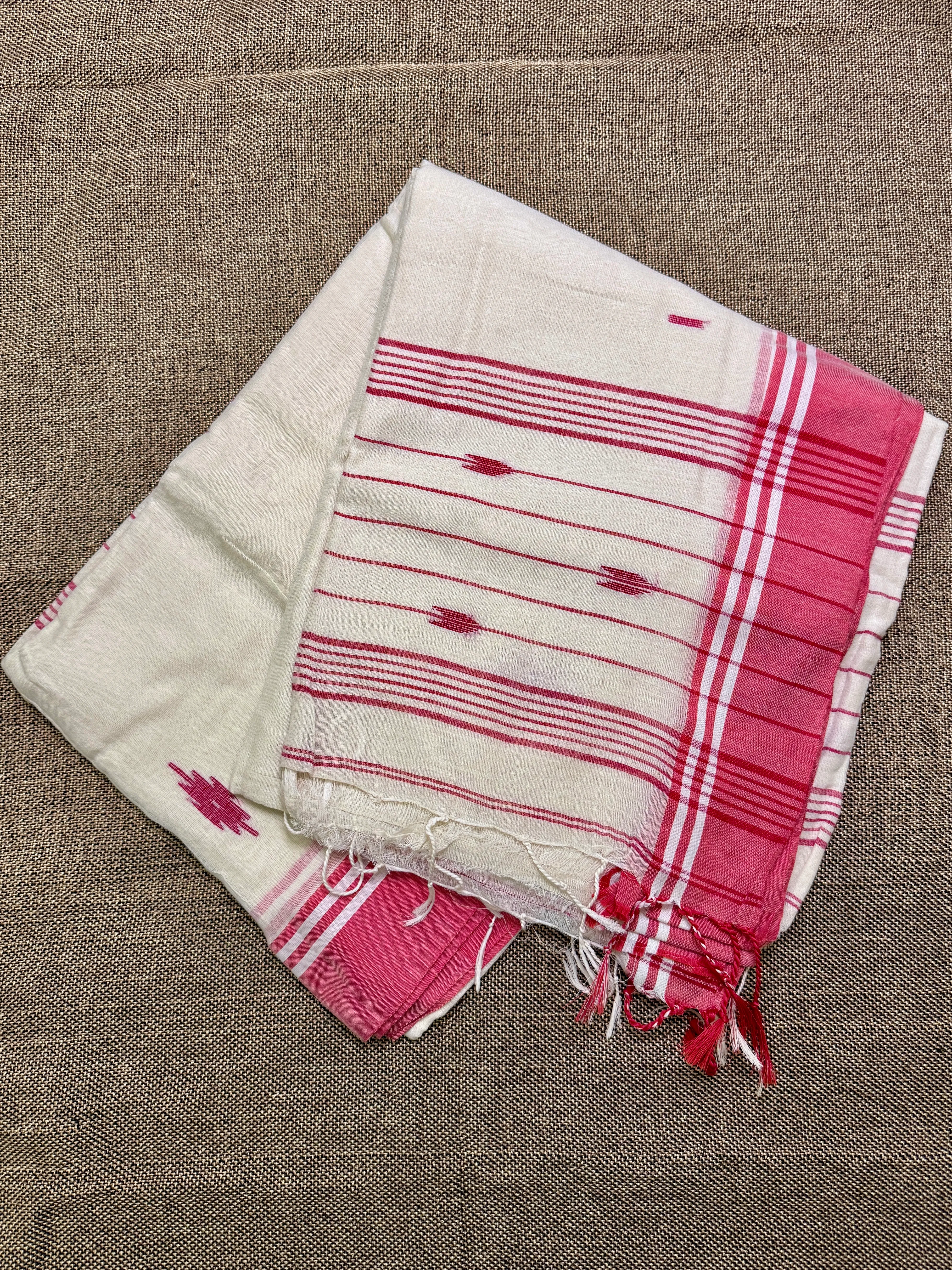 Folded cream mul cotton Tangail saree with fine red stripes and a pink plaid border, laid on a woven jute surface.